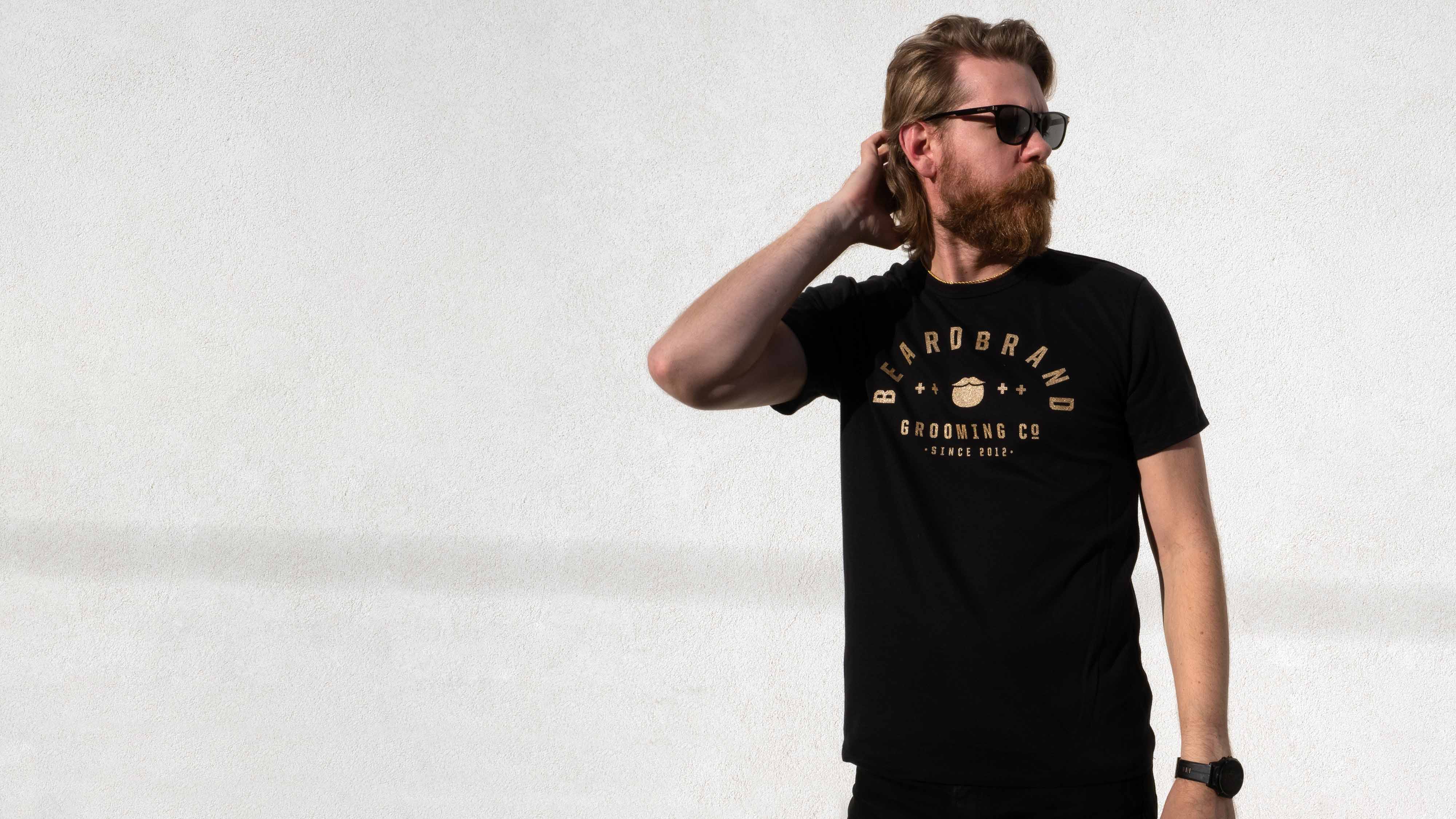 Eric Bandholz wearing dark sunglasses and a black Beardbrand Banner Tee in front of textured white wall. His right hand is raised to the nape of his neck and he has a dark watch on his left wrist.