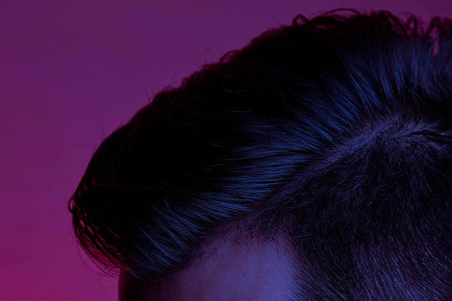 Styled hair focused on the top left of someone's head who is looking down, showing their side part close up in a gradient of magenta, purple and blue lighting.