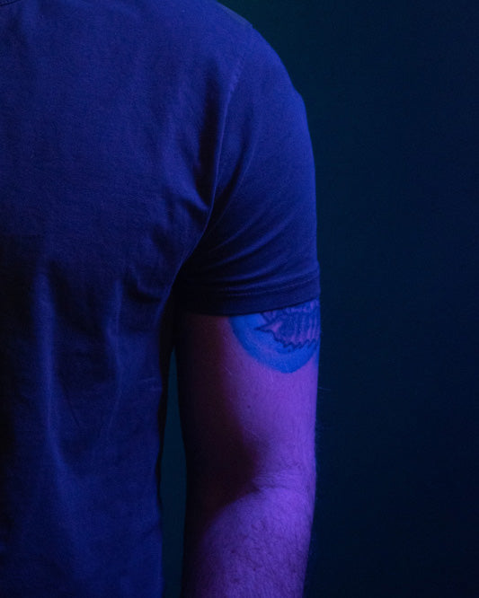 A person's torso and arm highlighted in blue and purple lighting. Part of a tattoo is visible below their navy blue t-shirt sleeve.