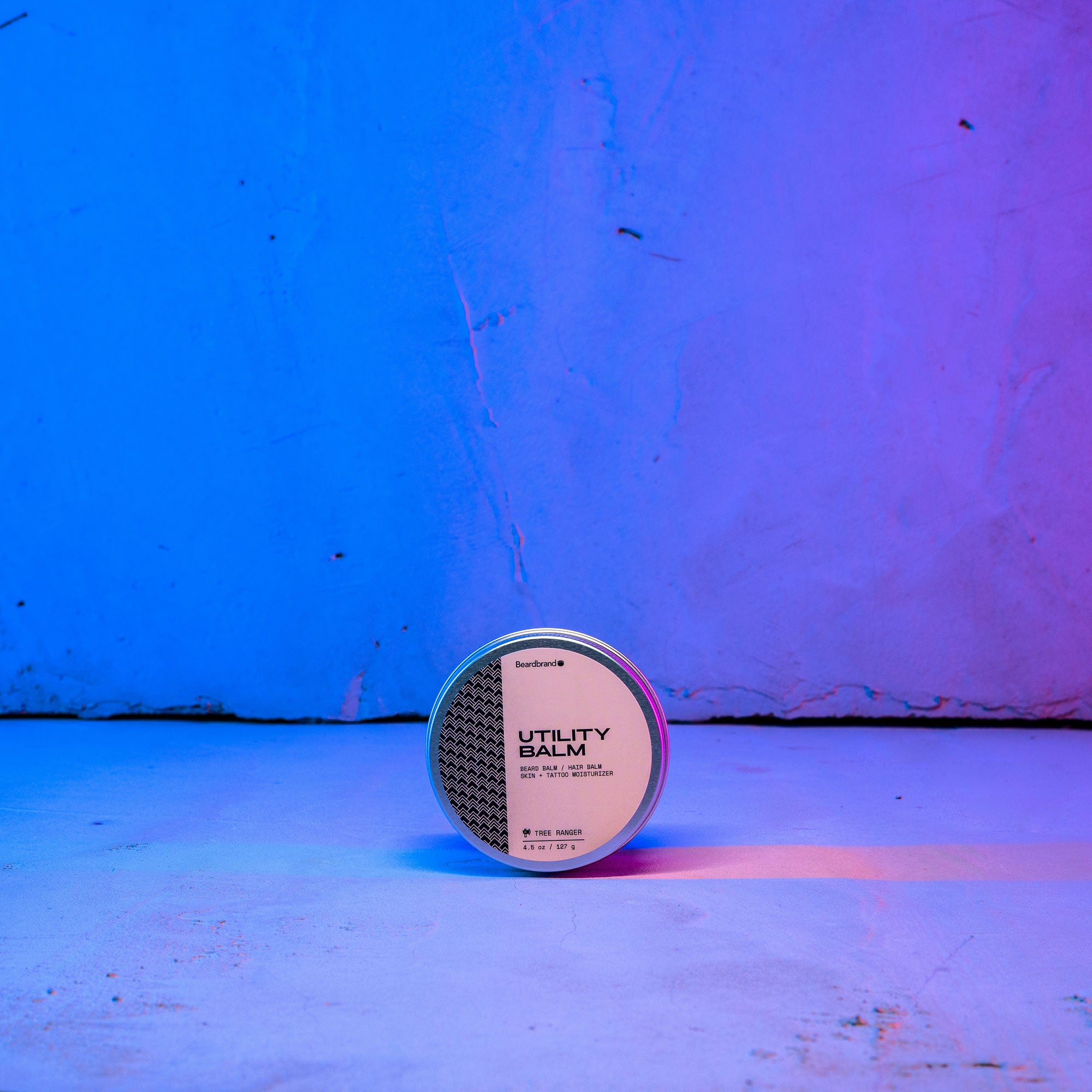 Beardbrand's Utility Balm set on a concrete backdrop highlighted in blue and pink lighting with the label facing forward.