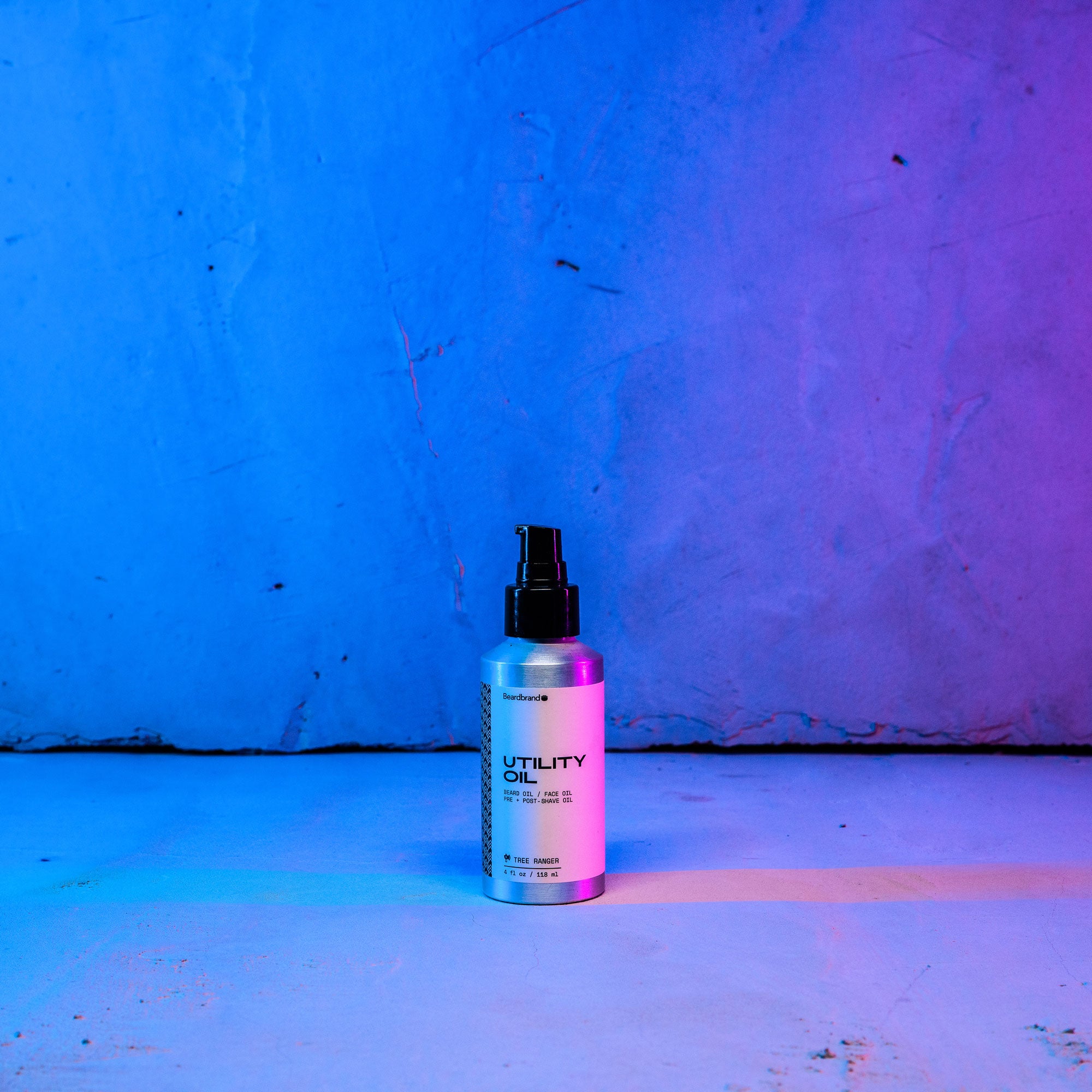 Beardbrand's Utility Oil set on a concrete backdrop highlighted in blue and pink lighting with the label facing forward.