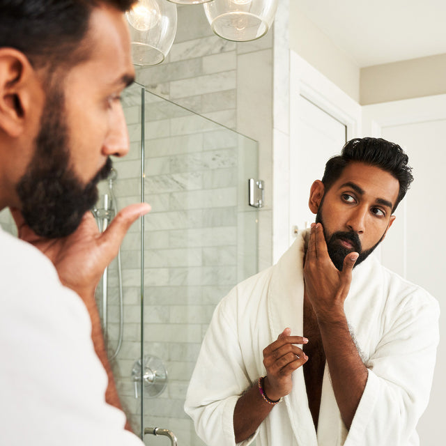 A beardsman wearing a robe getting ready in front of a mirror.