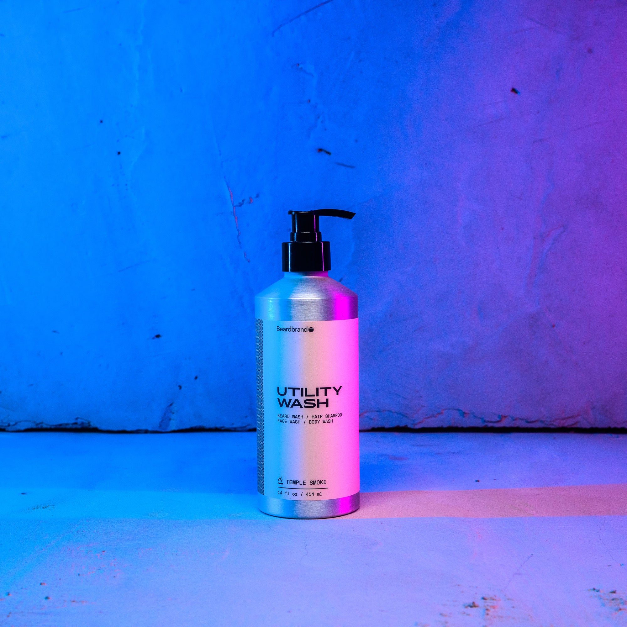 Beardbrand's Utility Wash set on a concrete backdrop highlighted in blue and pink lighting with the label facing forward.